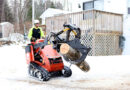 A tree worker uses a skid steer to carry a large log from a job site.